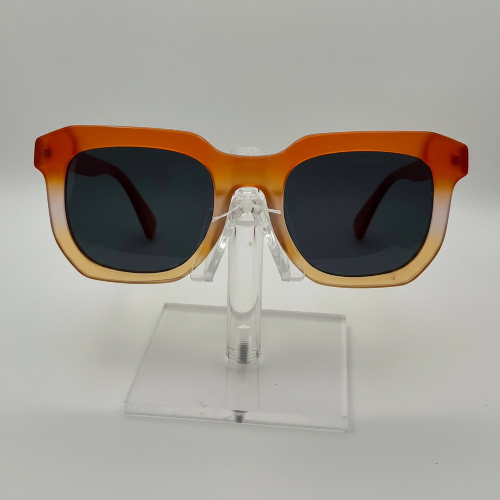 Square sunglasses with a translucent orange-to-tan gradient frame and dark lenses, displayed on a clear acrylic stand against a light gray background.