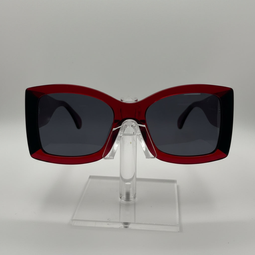 A pair of oversized square sunglasses with a translucent dark red frame and dark gray lenses, displayed on a clear acrylic stand against a light gray background.