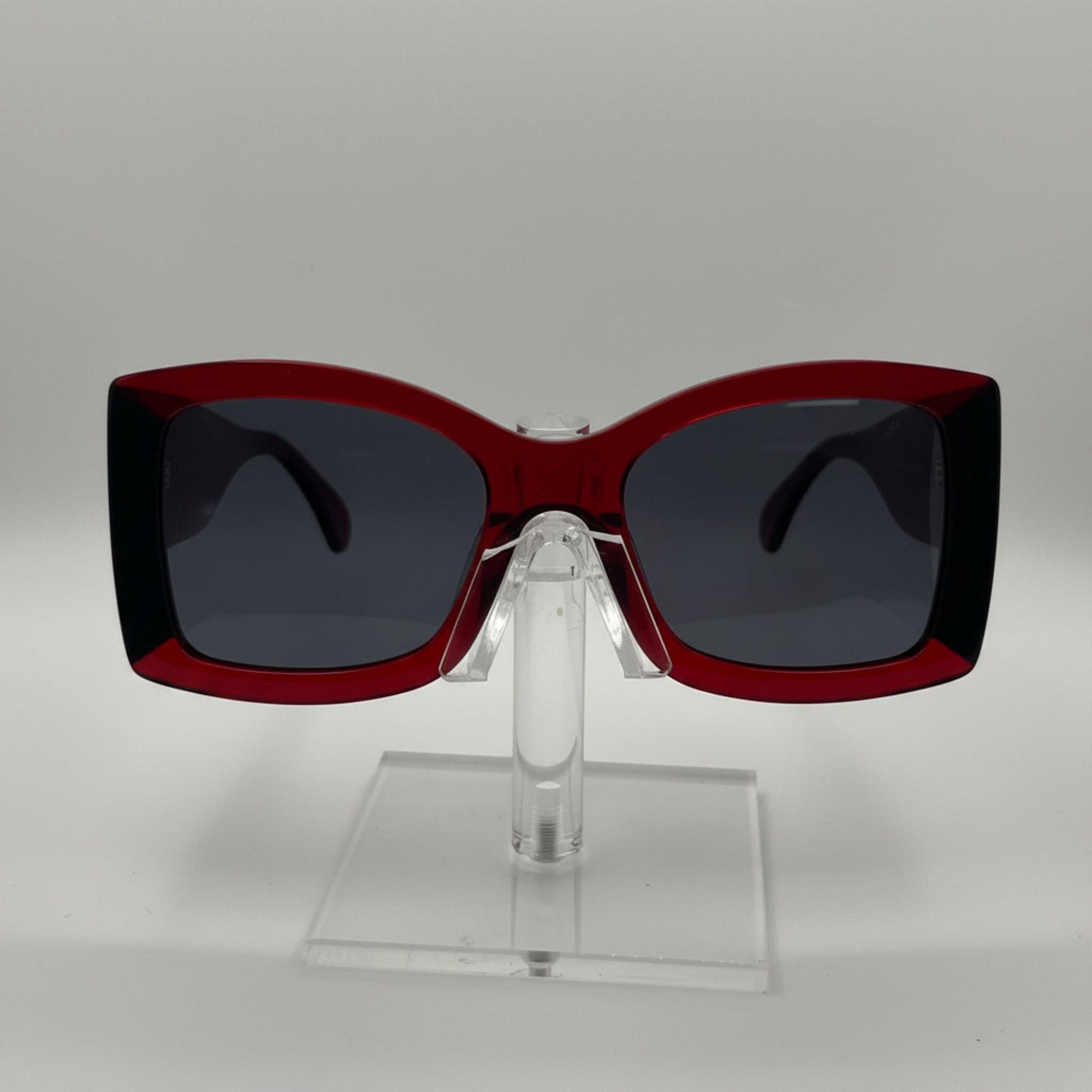 A pair of oversized square sunglasses with a translucent dark red frame and dark gray lenses, displayed on a clear acrylic stand against a light gray background.