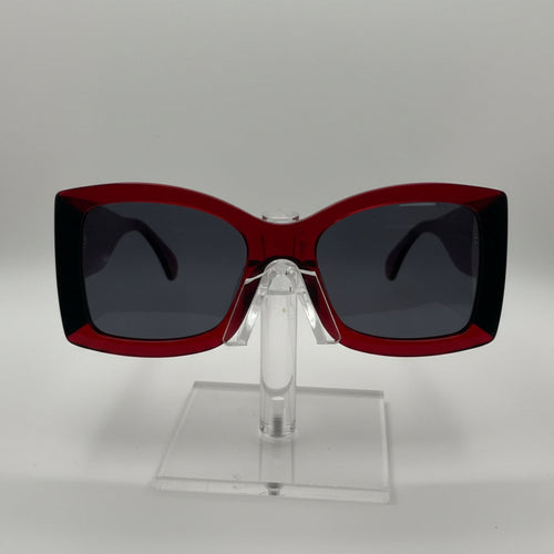A pair of oversized square sunglasses with a translucent dark red frame and dark gray lenses, displayed on a clear acrylic stand against a light gray background.