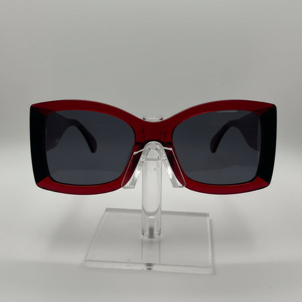 A pair of oversized square sunglasses with a translucent dark red frame and dark gray lenses, displayed on a clear acrylic stand against a light gray background.