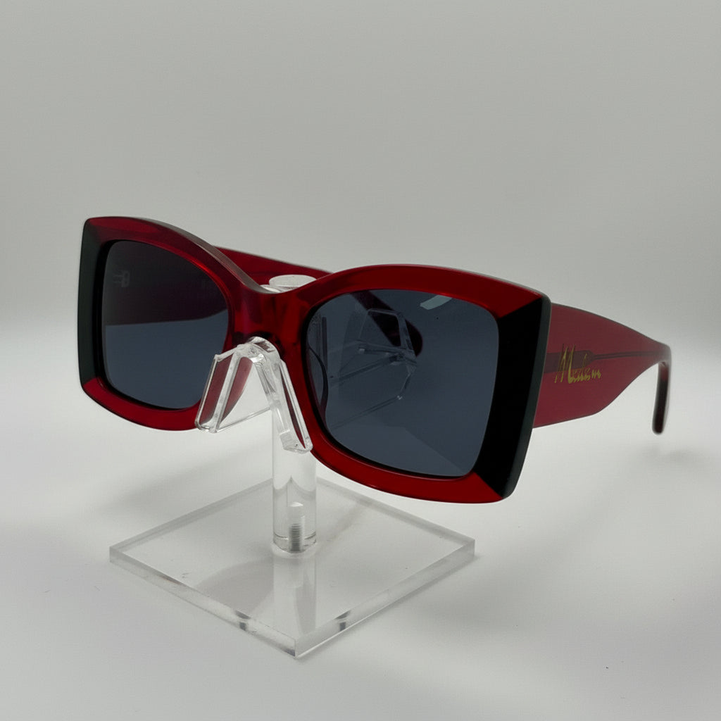A pair of square sunglasses with a translucent dark red acetate frame, dark lenses, and a gold script logo on the temple, displayed on a clear acrylic stand against a light gray background.