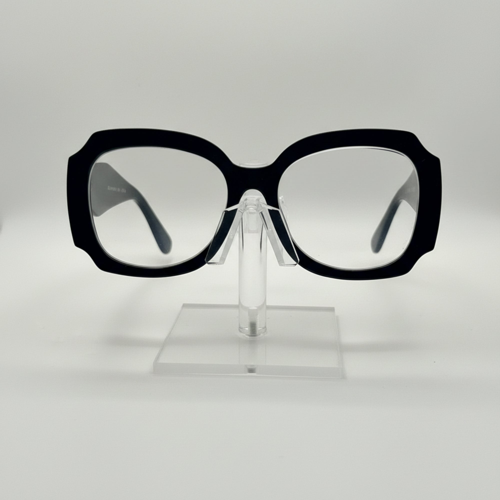 Oversized square eyeglasses with a thick black frame and clear lenses, displayed on a transparent acrylic stand against a light gray background.