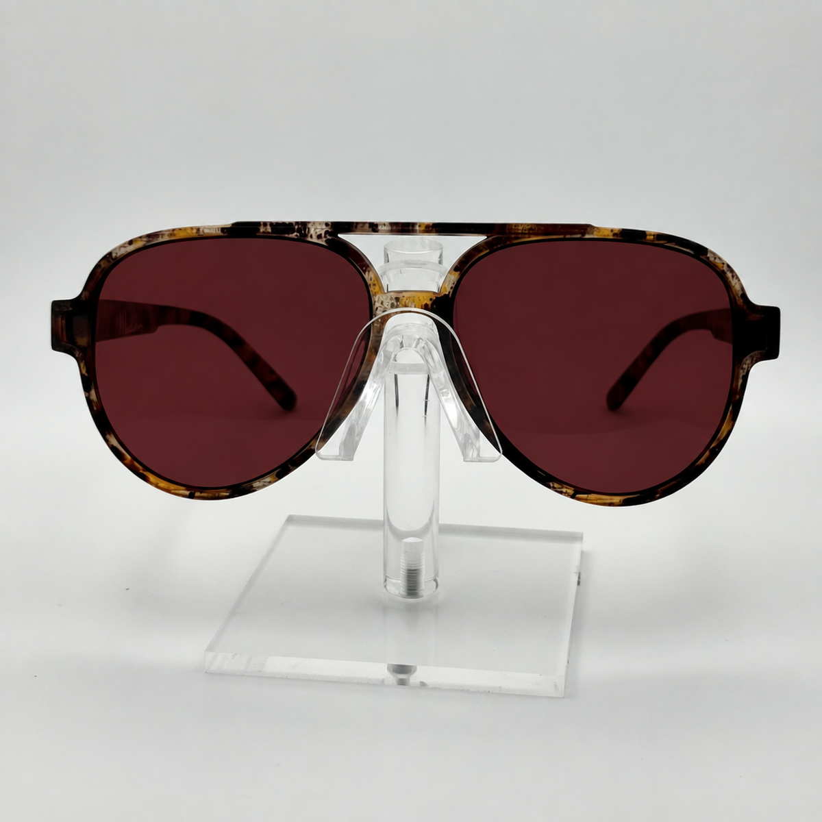 A pair of aviator-style sunglasses with a tortoiseshell frame and dark reddish-brown lenses, displayed on a clear acrylic stand against a light gray background.
