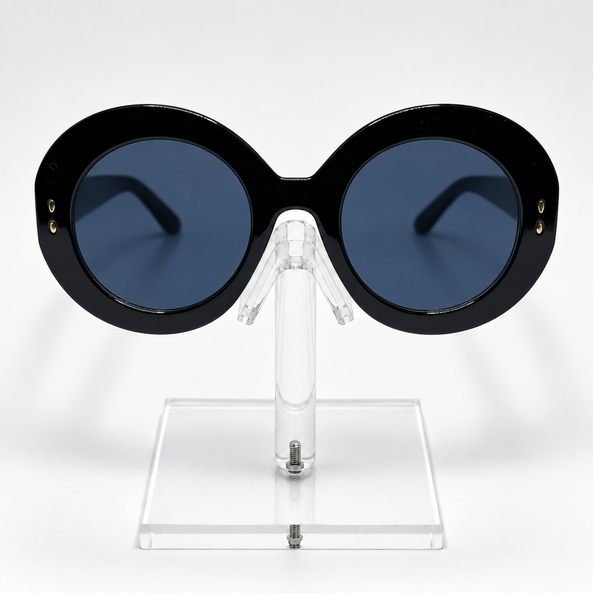 Oversized round sunglasses with a thick black frame and dark blue lenses, displayed on a clear acrylic stand against a white background.