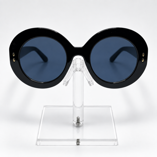 Oversized round sunglasses with a thick black frame and dark blue lenses, displayed on a clear acrylic stand against a white background.