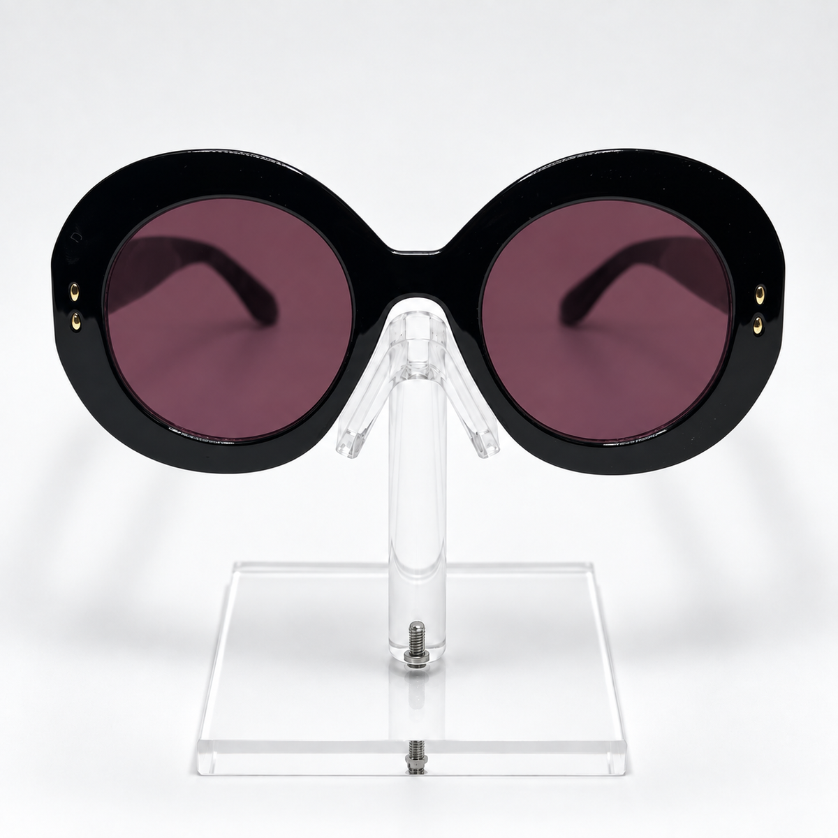 Oversized round sunglasses with thick black frames and dark pinkish-purple lenses, displayed on a clear acrylic stand against a white background.
