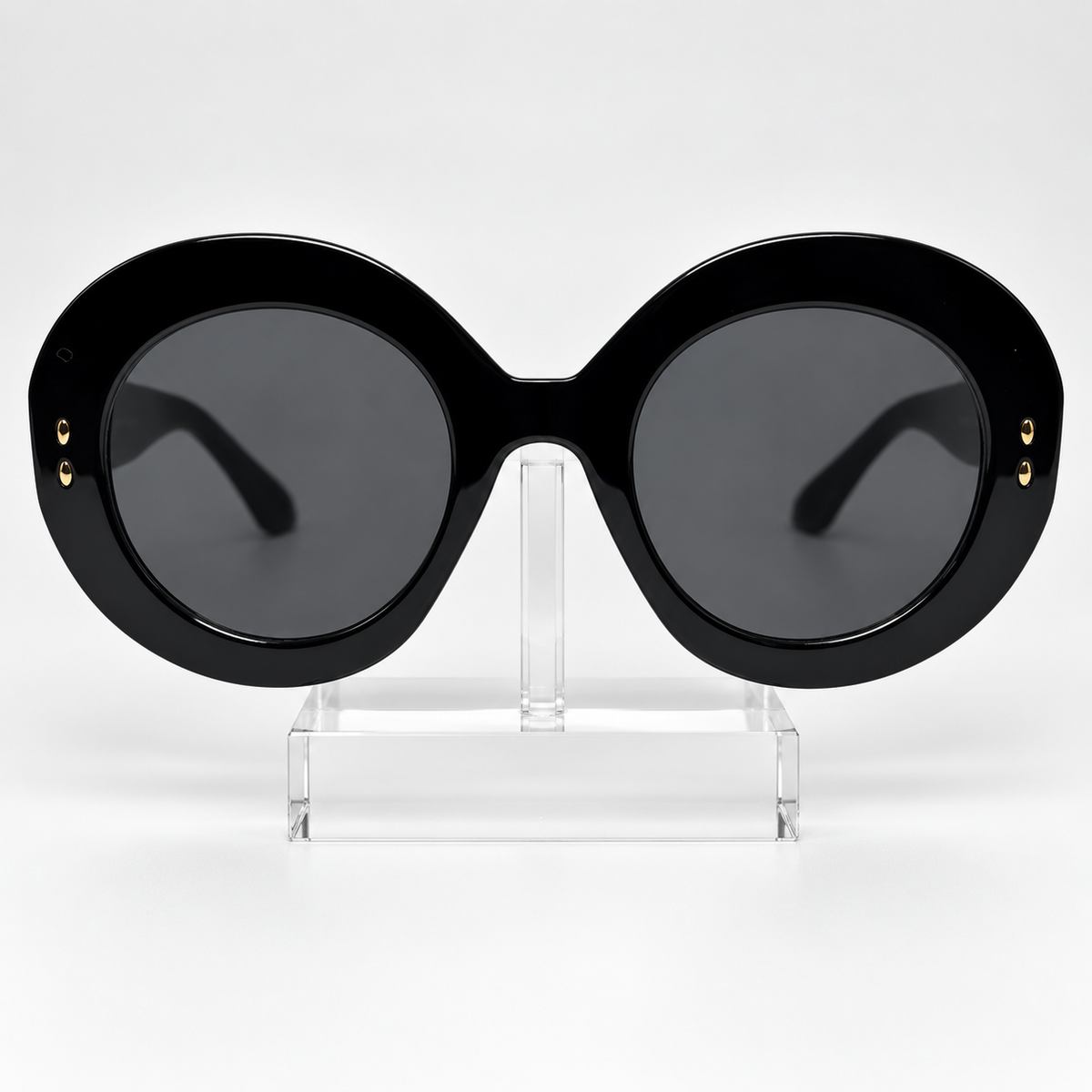 Oversized round sunglasses with a thick black frame and dark lenses, displayed on a clear acrylic stand against a light gray background.