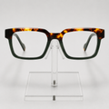 Rectangular eyeglasses with a dark green and amber tortoiseshell acetate frame, displayed on a clear acrylic stand against a light gray background.