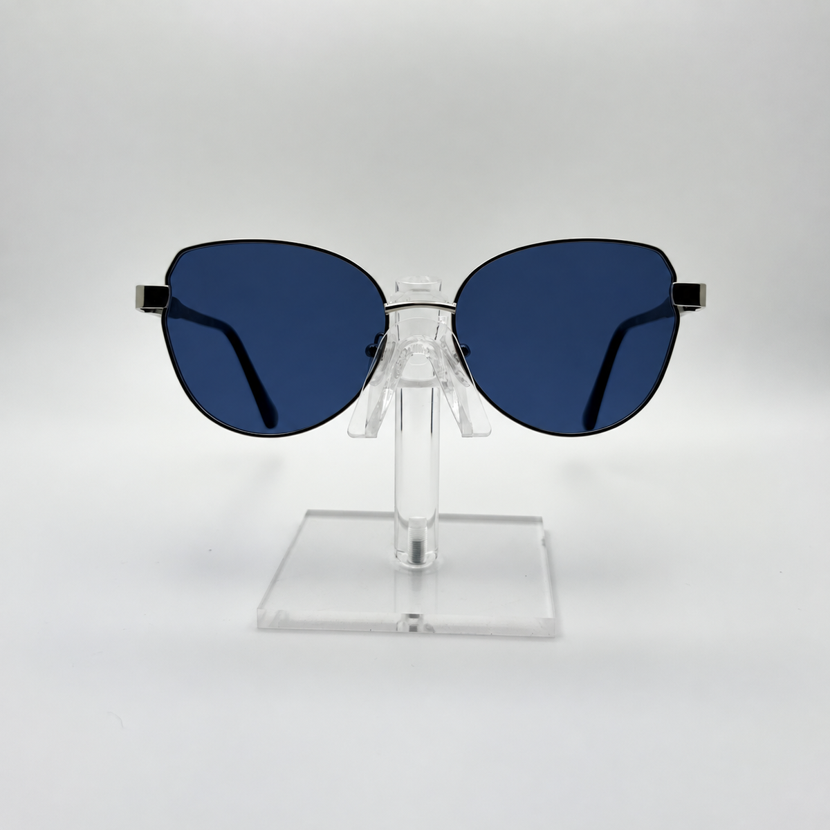 Oversized cat-eye sunglasses with dark blue lenses and a thin black metal frame, displayed on a clear acrylic stand against a light gray background.