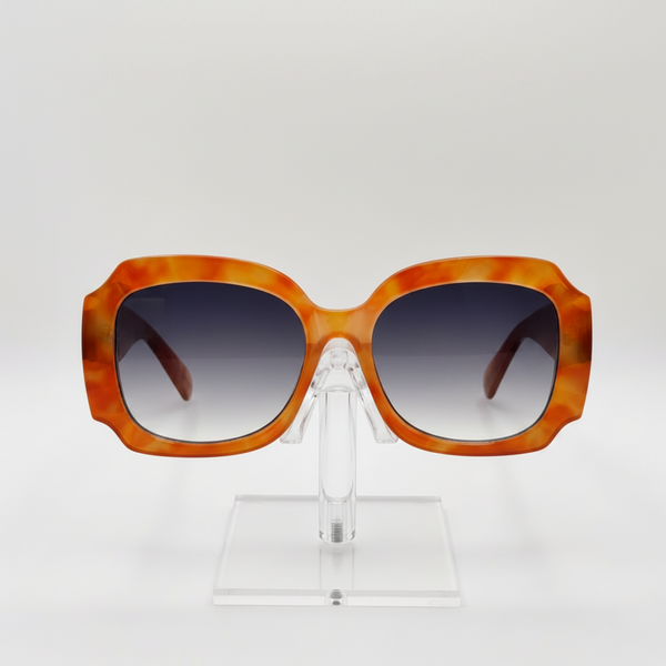 Oversized square sunglasses with a translucent orange and yellow marbled acetate frame and dark gradient lenses, displayed on a clear acrylic stand against a light gray background.
