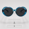 Oval sunglasses with translucent blue frames and dark gray lenses, displayed on a clear acrylic stand against a light gray background.