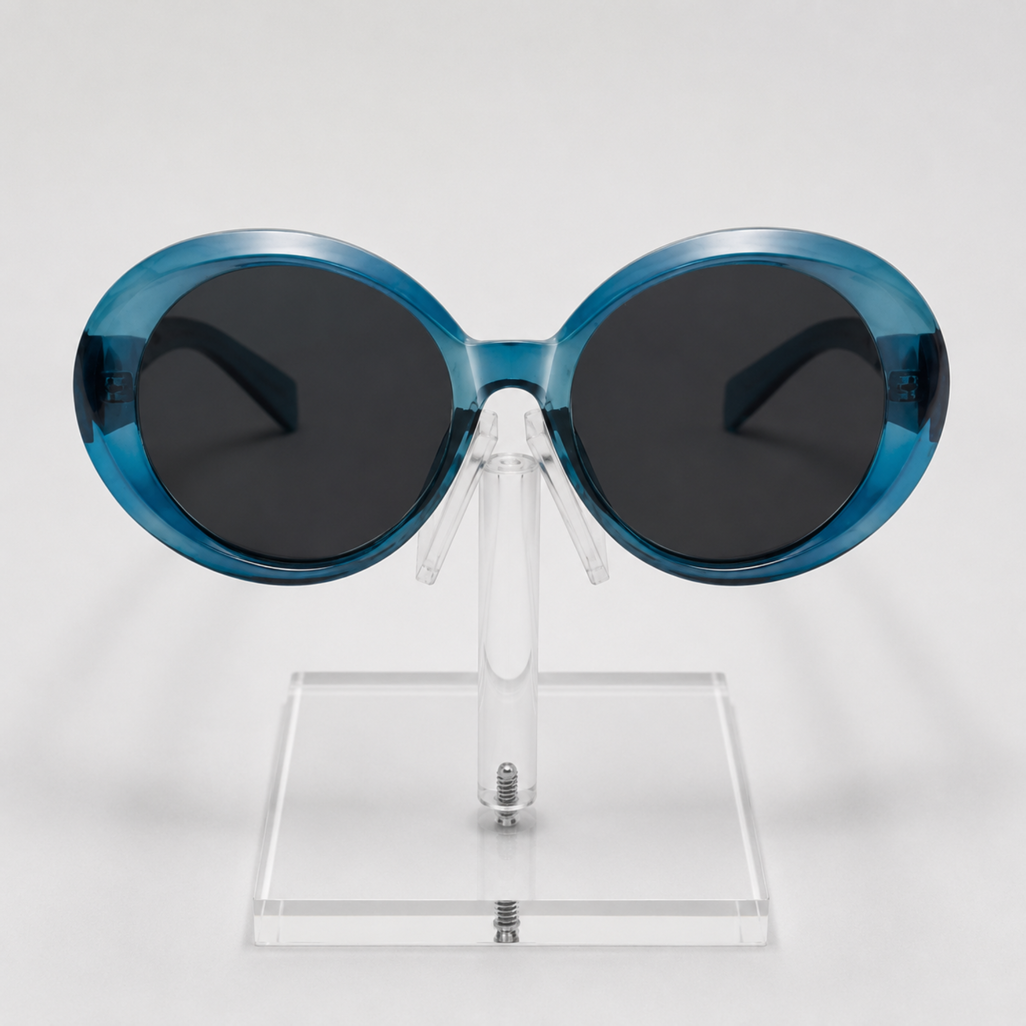 Oval sunglasses with translucent blue frames and dark gray lenses, displayed on a clear acrylic stand against a light gray background.