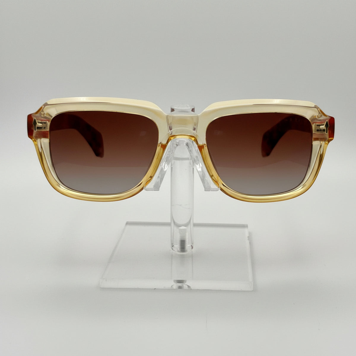 Gold-framed sunglasses with brown lenses on a clear stand against a gray background