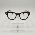 A pair of classic-style eyeglasses with a dark brown and black tortoiseshell acetate frame, displayed on a clear acrylic stand against a light gray background.