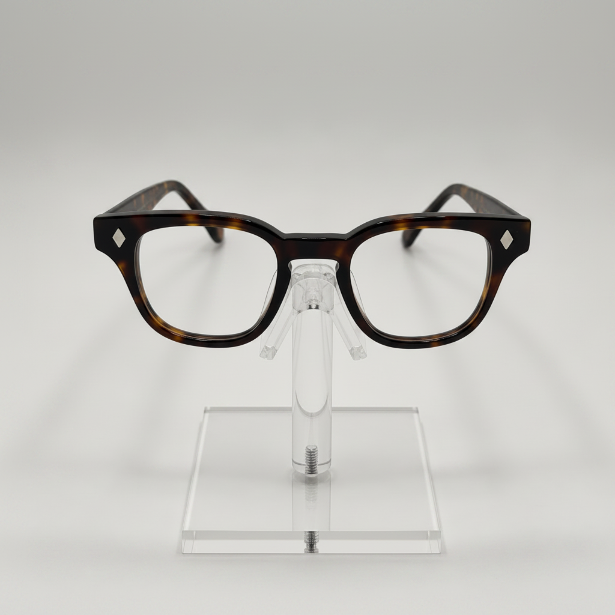A pair of classic-style eyeglasses with a dark brown and black tortoiseshell acetate frame, displayed on a clear acrylic stand against a light gray background.