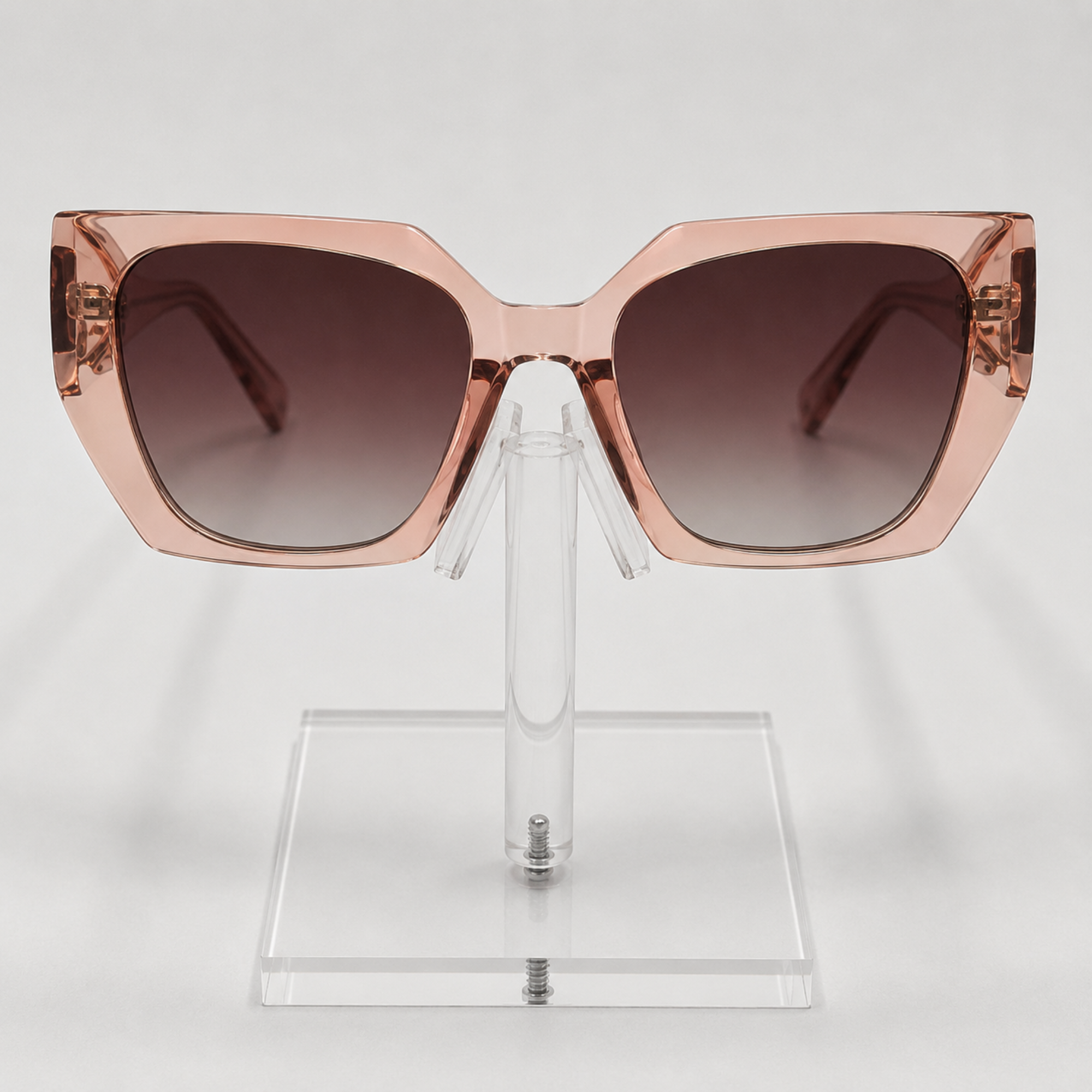 Oversized square sunglasses with a translucent rose gold frame and dark brown gradient lenses, displayed on a clear acrylic stand against a light gray background.