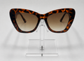 Tortoiseshell cat-eye sunglasses with brown gradient lenses, displayed on a clear acrylic stand against a light gray background.