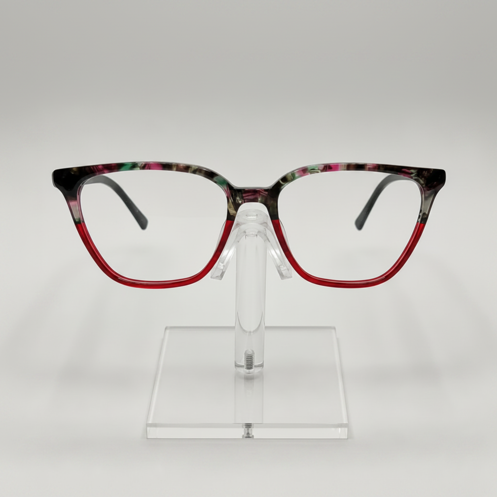 Fashionable square eyeglasses with a red acetate lower frame and a black, green, and pink marbled top frame, displayed on a clear stand against a light gray background.