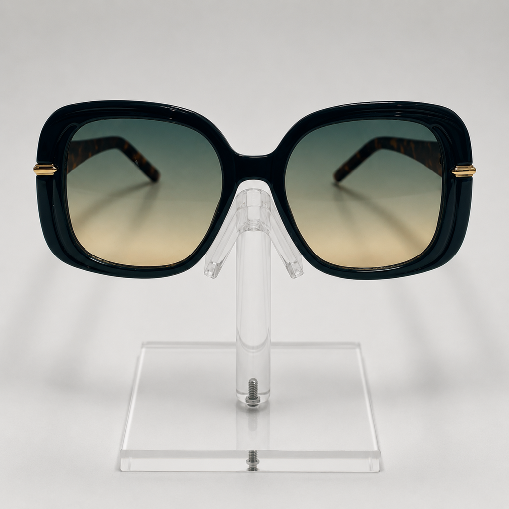 Oversized square sunglasses with a glossy black frame, gradient lenses that fade from dark green to beige, and gold metal accents on the temples, displayed on a clear acrylic stand.