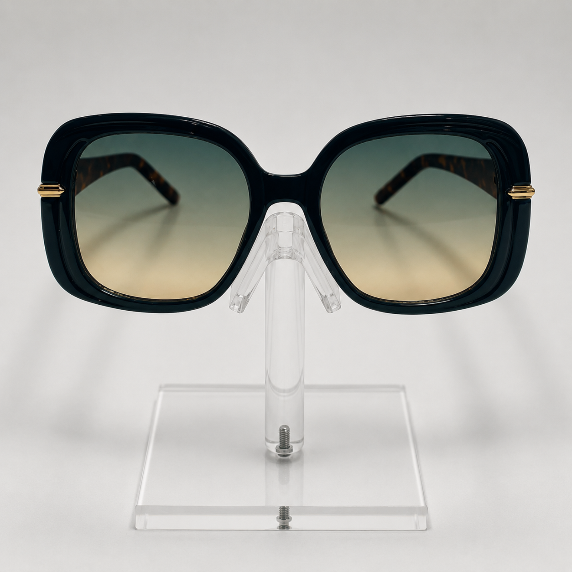 Oversized square sunglasses with a glossy black frame, gradient lenses that fade from dark green to beige, and gold metal accents on the temples, displayed on a clear acrylic stand.