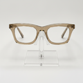 A pair of chunky, oversized square eyeglass frames in a translucent warm taupe-brown acetate, displayed on a clear acrylic stand against a clean white background.