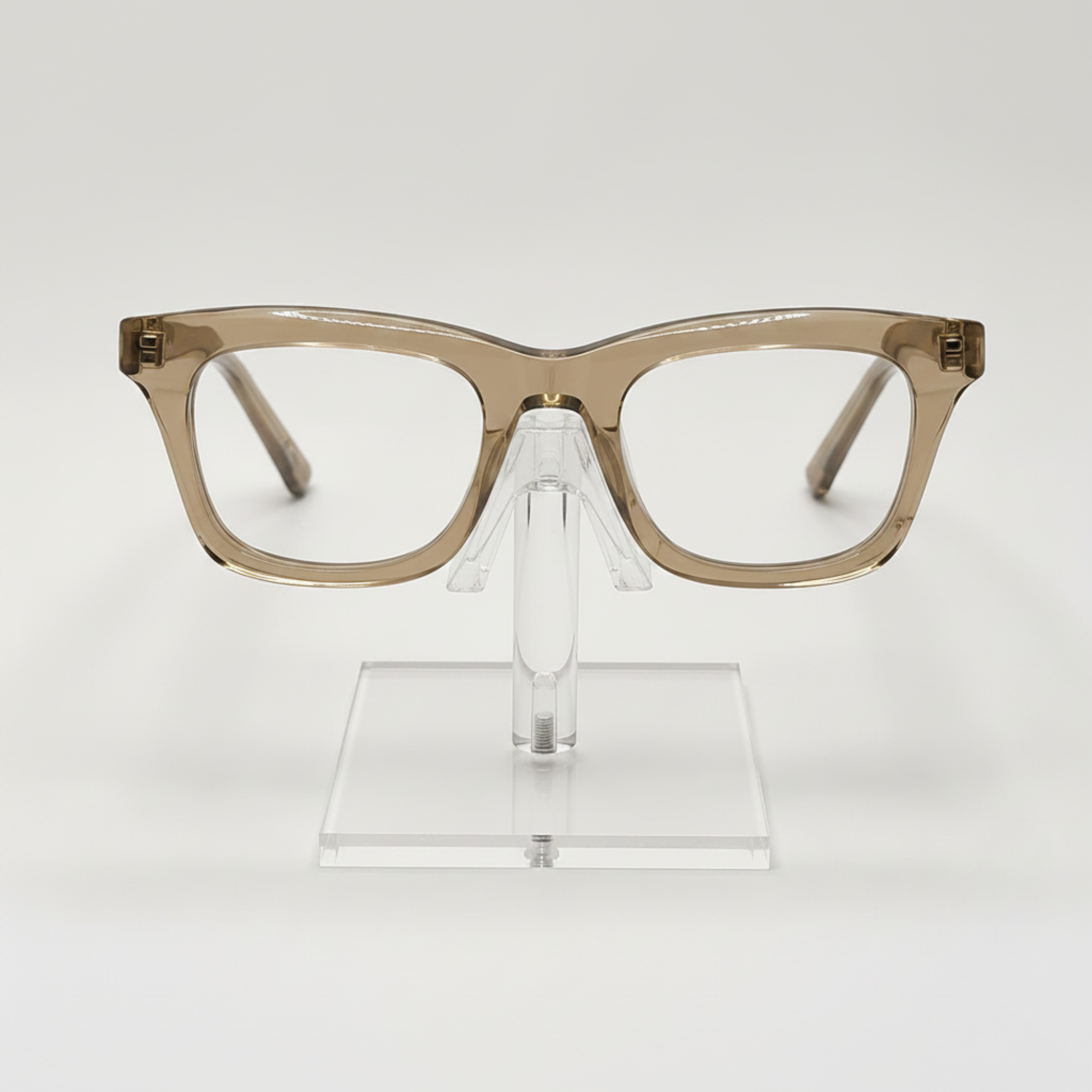 A pair of chunky, oversized square eyeglass frames in a translucent warm taupe-brown acetate, displayed on a clear acrylic stand against a clean white background.