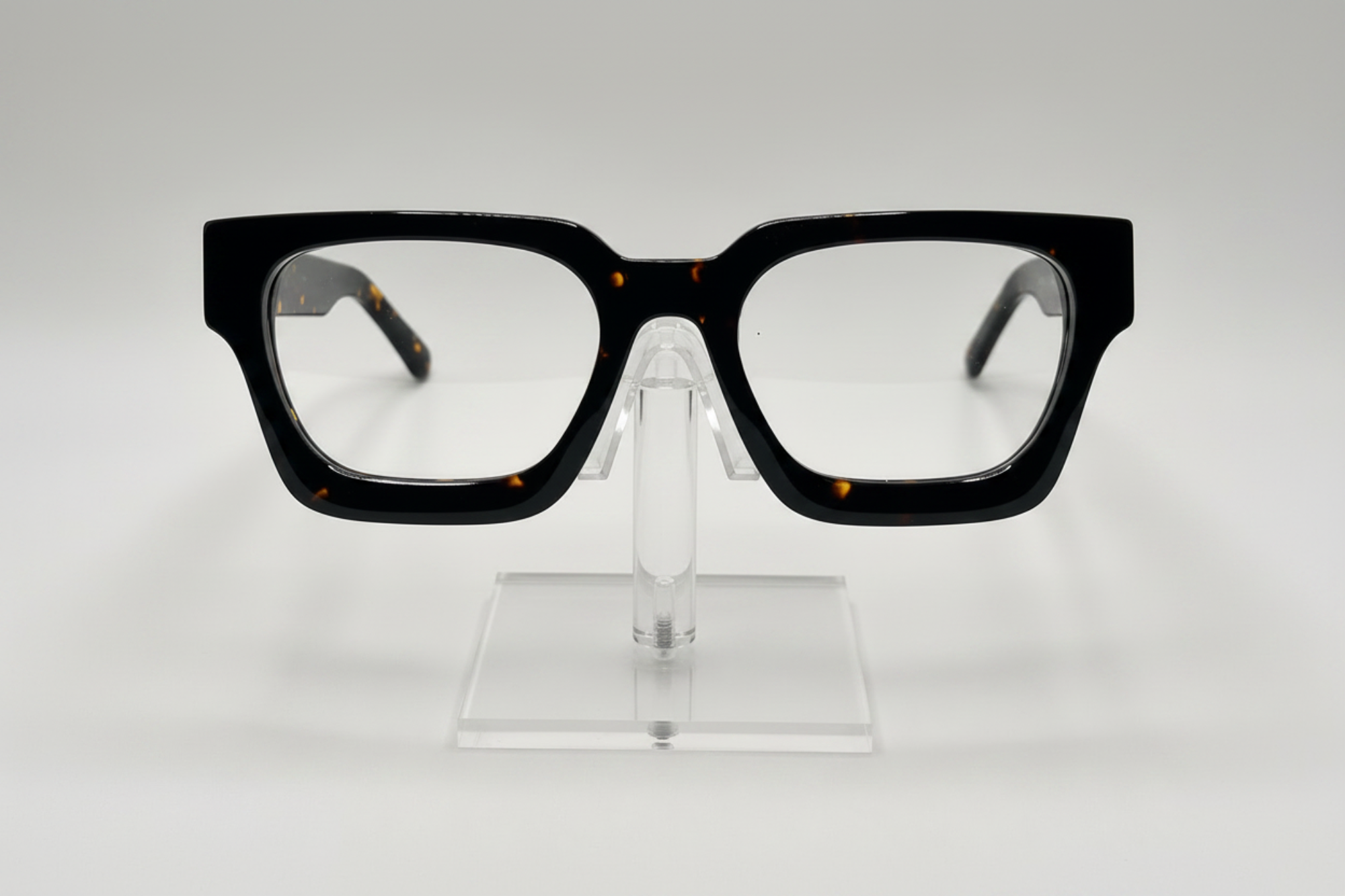 Black square eyeglasses with a subtle tortoiseshell pattern on the inner temples, displayed on a clear acrylic stand against a light gray background.