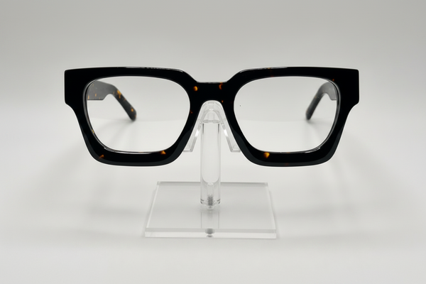 Black square eyeglasses with a subtle tortoiseshell pattern on the inner temples, displayed on a clear acrylic stand against a light gray background.