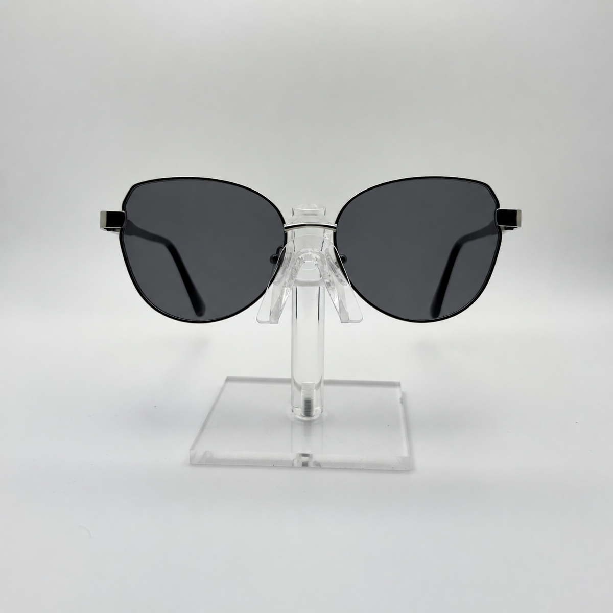 Oversized cat-eye sunglasses with dark gray lenses and a thin black metal frame, displayed on a clear acrylic stand against a light gray background.