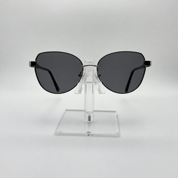 Oversized cat-eye sunglasses with dark gray lenses and a thin black metal frame, displayed on a clear acrylic stand against a light gray background.