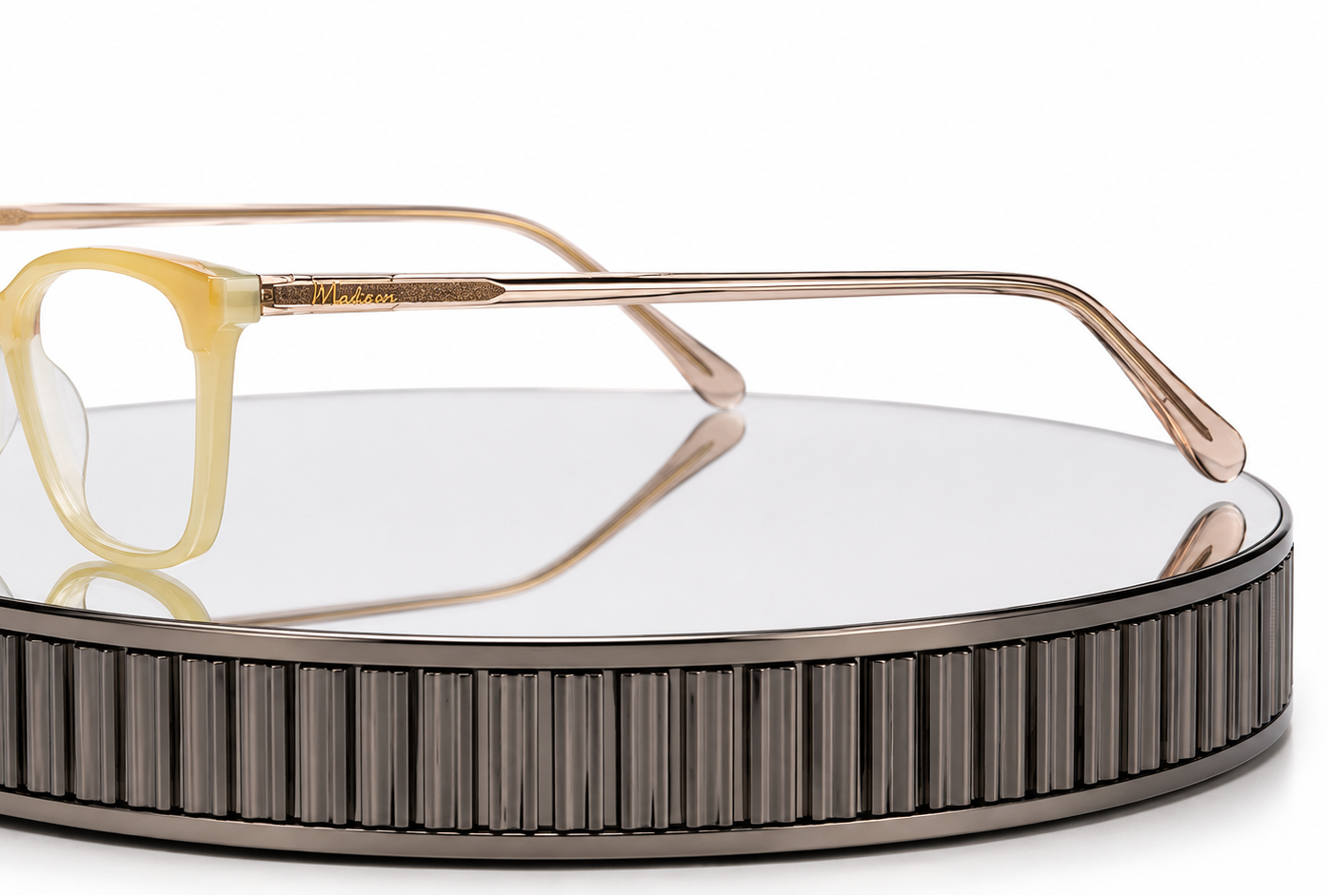 A pair of eyeglasses with translucent pale yellow acetate frames and rose gold metal temples, displayed on a round mirrored stand with a ribbed dark metallic base against a white background.